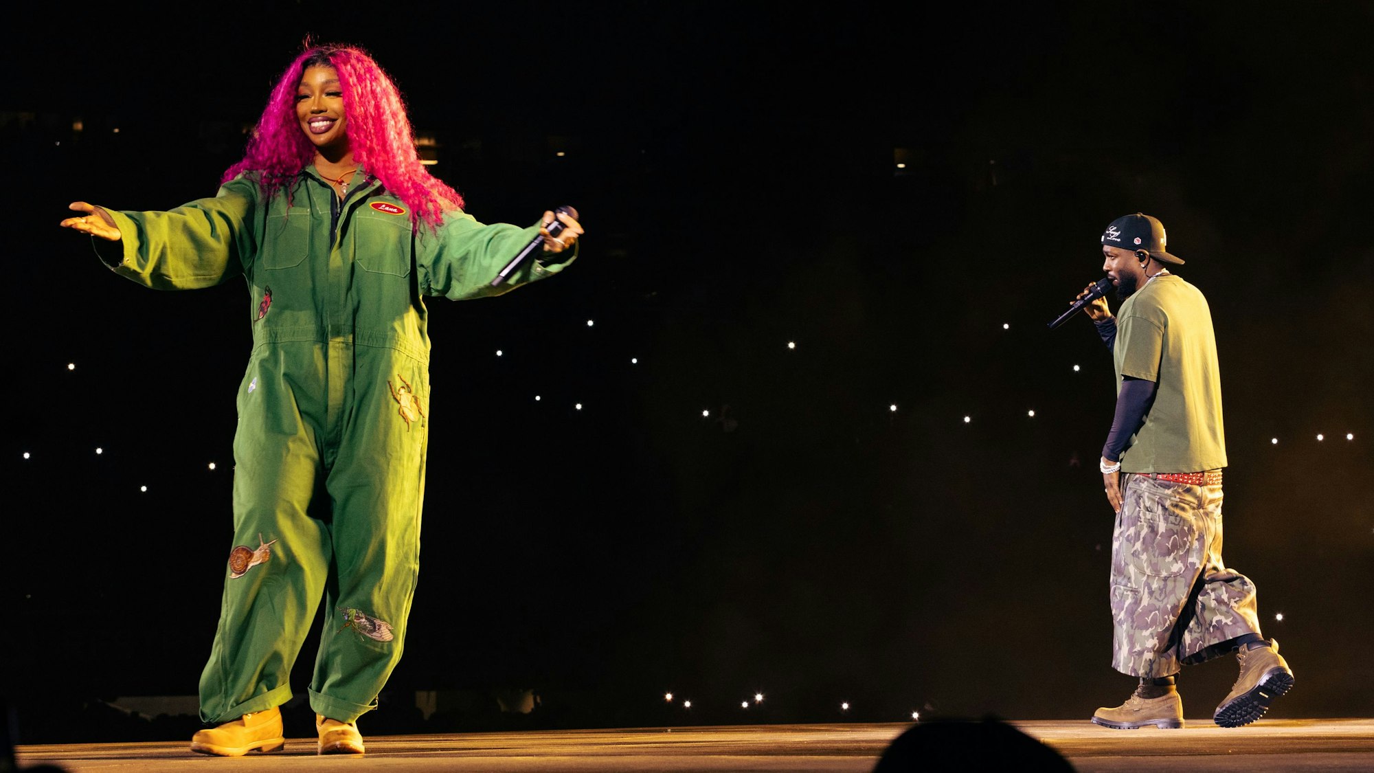 SZA steht links im grünen Overall auf dem Laufsteg im Stadioninnenraum, Kendrick Lamar rappt, von der Kamera abgewandt, rechts im Bild.