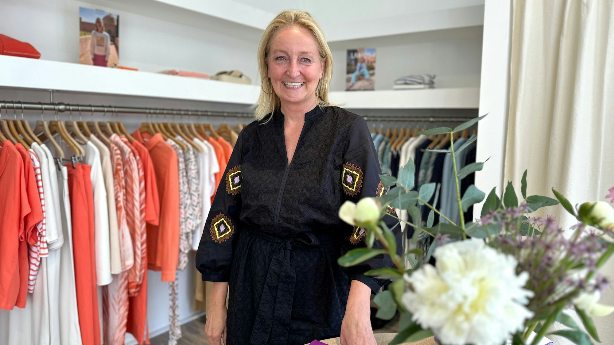 Eine Frau mit blondem schulterlangem Haar und einer schwarzen Bluse steht in einem Modegeschäft.