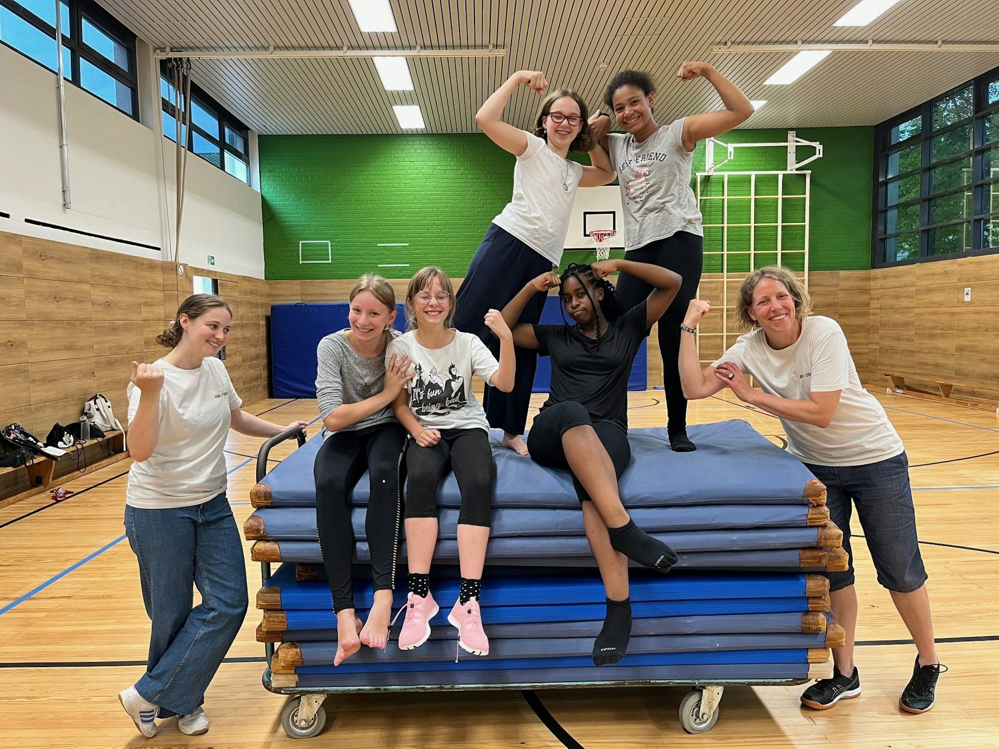 Fünf Teilnehmerinnen der „Starke Mädchen“-Gruppe sitzen auf einem Sport-Matten-Wagen, daneben stehen ihre Trainerinnen des Kölner Vereins „FairStärken e.V.“ Anne Arenhövel und Juliane Götz.