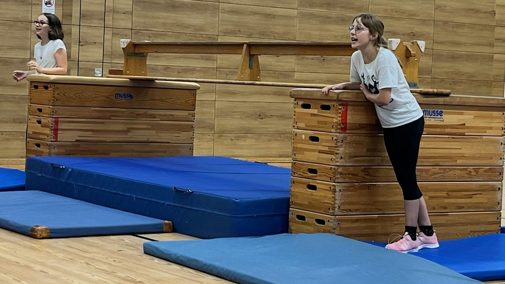 Zwei Teilnehmerinnen des „Starke Mädchen“-Projekts stehen neben zwei Holzkästen in einer Turnhalle und feuern ihre Mitstreiterinnen an.