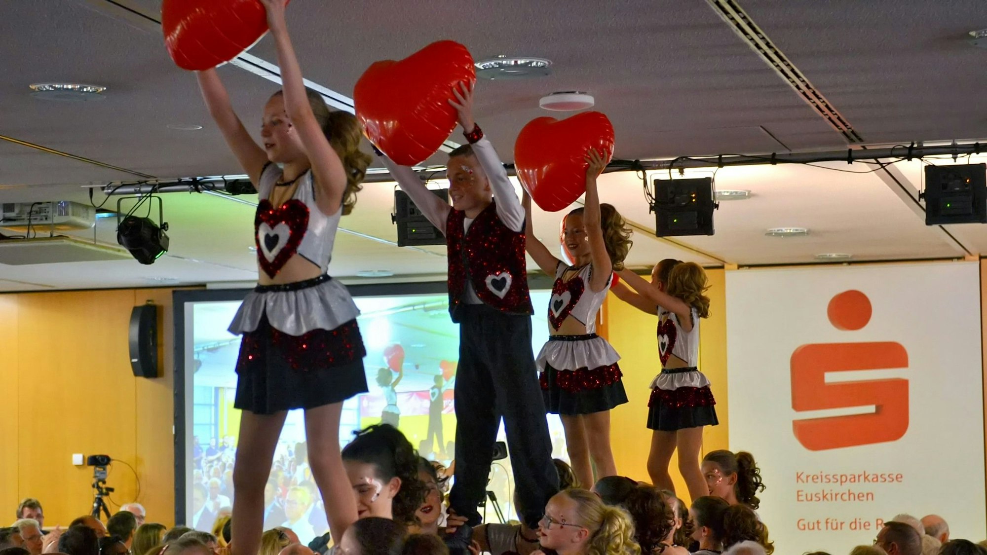 Drei Mädchen und ein Junge stehen auf den Schultern von weiteren Kindern und halten Luftballon-Herzen hoch vor sich.