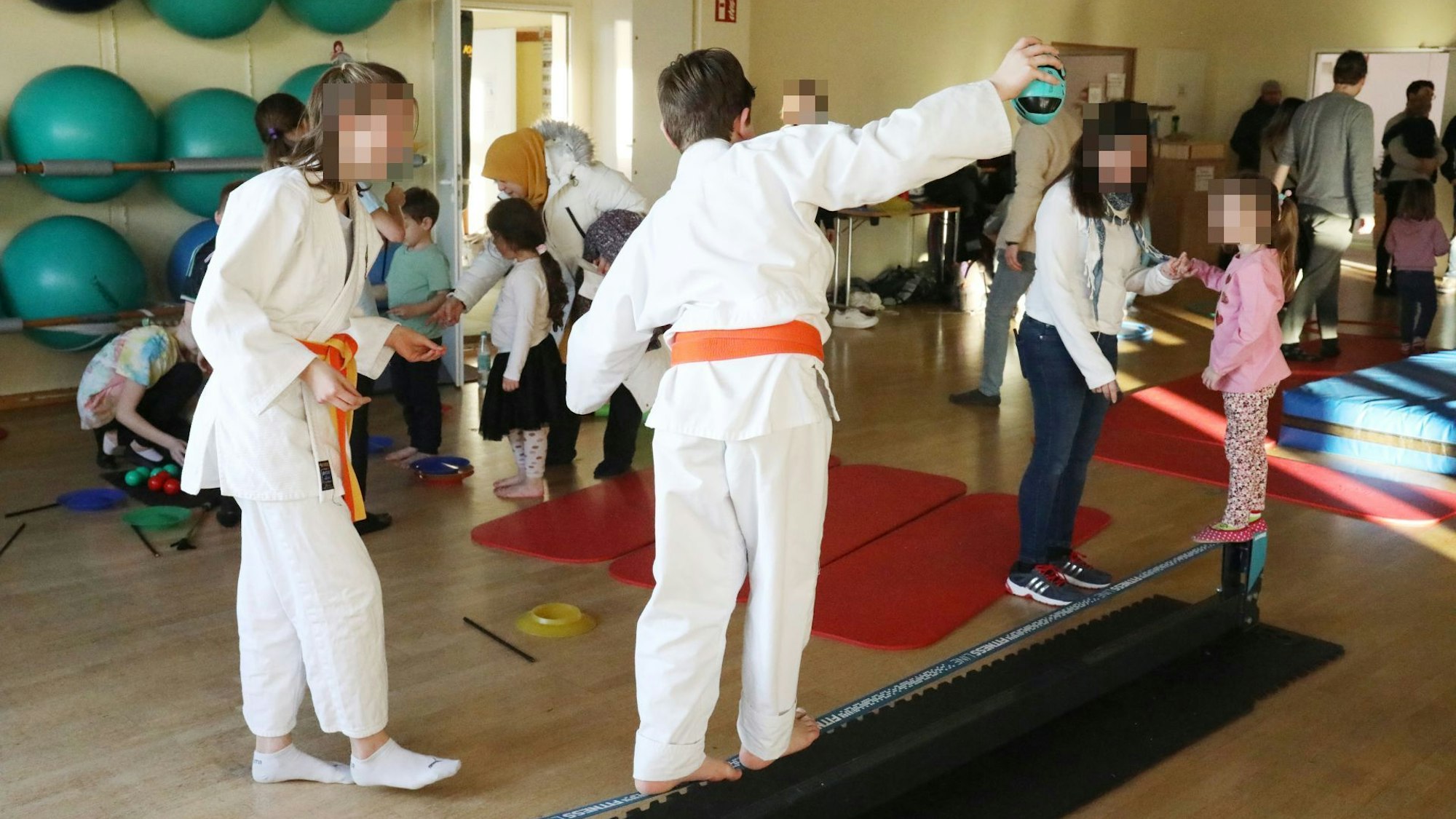 Kinder und Jugendliche in einem Raum mit Sportgeräten. Zwei von ihnen tragen Judo-Kleidung.
