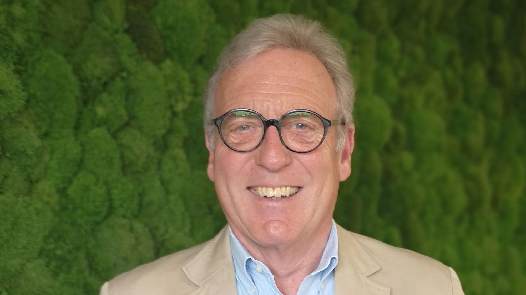 Auf dem Foto ist ein Mann mit Brille und grauem Haar zu sehen. Er trägt ein hellblaues Hemd, darüber ein beigefarbenes Sakko.