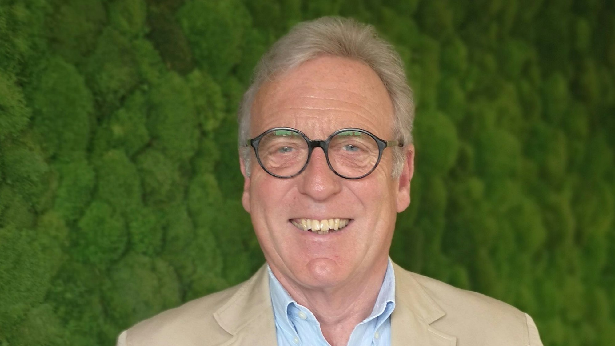 Auf dem Foto ist ein Mann mit Brille und grauem Haar zu sehen. Er trägt ein hellblaues Hemd, darüber ein beigefarbenes Sakko.