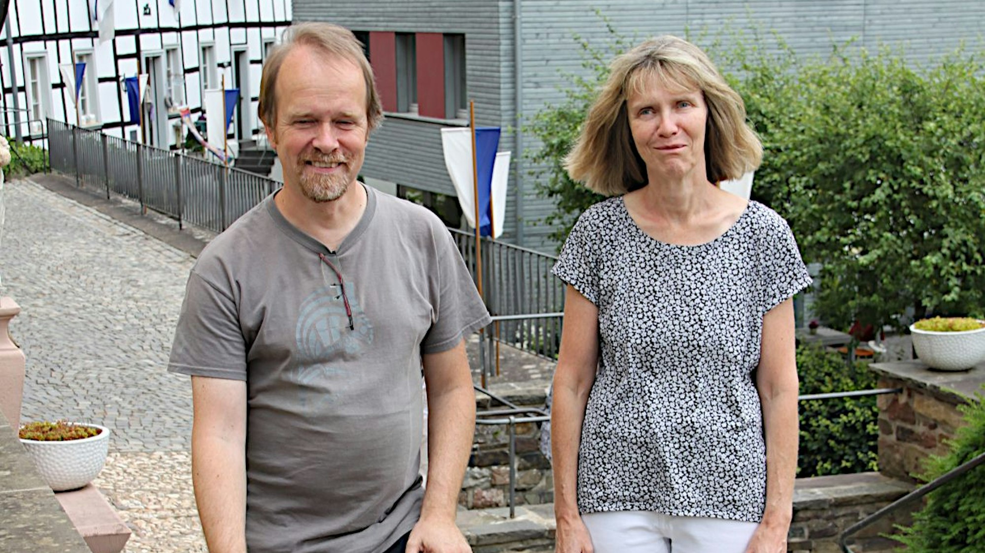 Ein Mann und eine Frau stehen nebeneinander, im Hintergrund ist ein mit Fahnen geschmücktes Fachwerkhaus zu sehen.