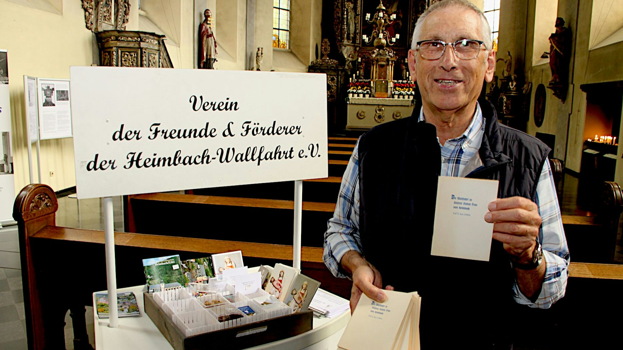 Ein Mann in blau-kariertem Hemd und dunkler Weste steht neben einem kleinen Verkaufsstand und hält ein Heft in die Kamera.