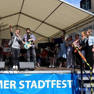 Auf der Bühne: Ein Musiker am Keyboard, einer an der Gitarre, einer mit einem Mikro, an der Seite rechts stehen mehrere Sängerinnen.