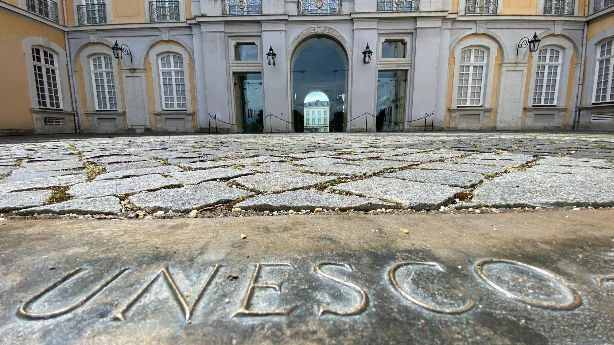 Zu sehen sind eine Bronzeplatte mit dem Schriftzug „Unesco“ und im Hintergrund das Schloss.