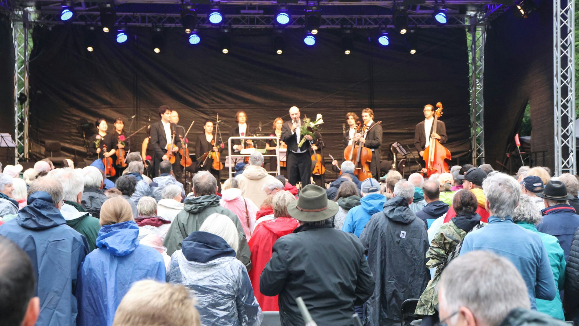 Ein Orchester auf der Bühne, davor Zuhörer in Regencapes.