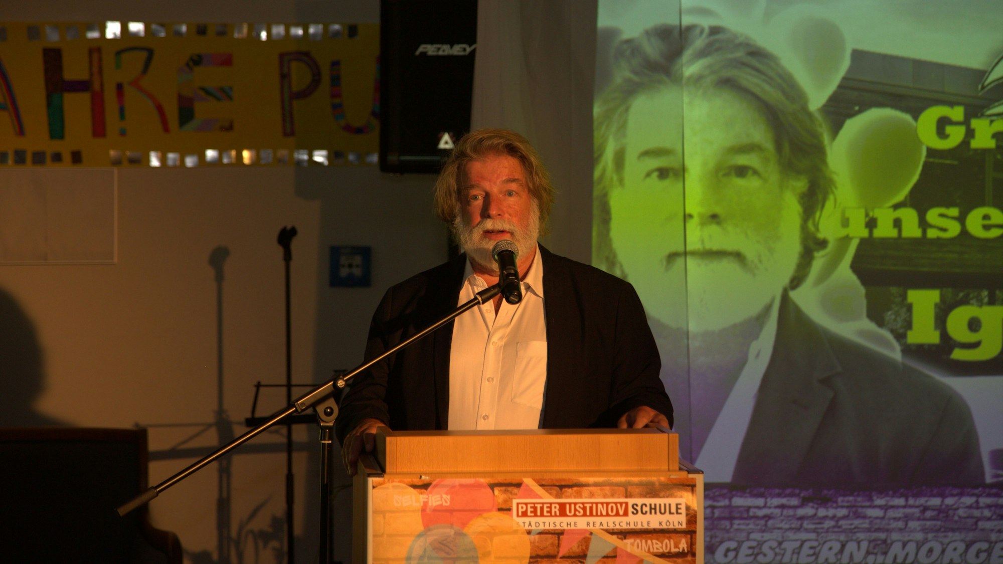 Ein Mann mit grauem Haar und weißem Bart steht an einem Pult und spricht in ein Mikrofon.