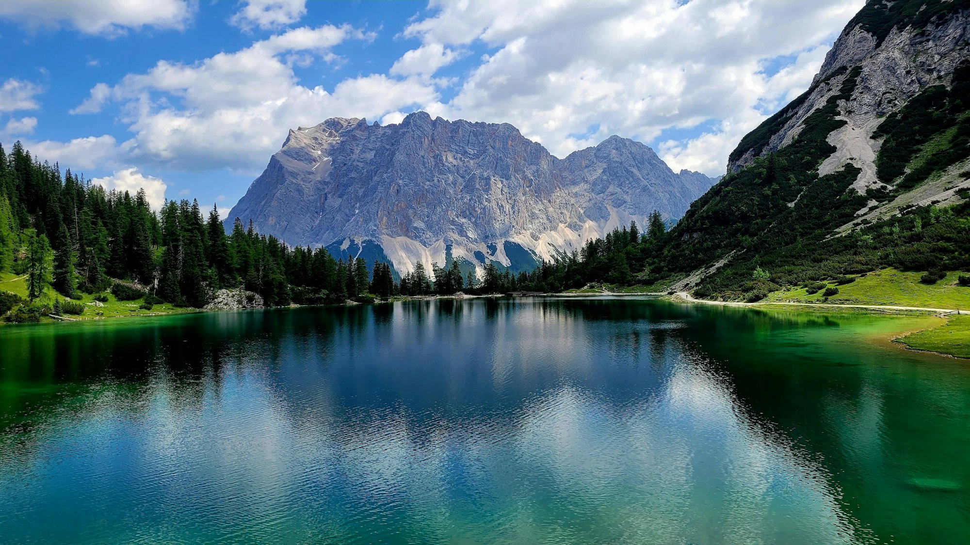 Blick auf den türkisblauen, von Bäumen eingerahmten Seebensee, auf dem Wanderweg zur Zugspitze. Im Hintergrund sind Berge zu sehen.