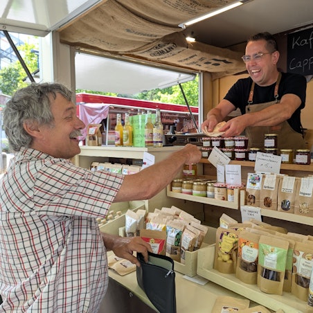 Das Foto zeigt den Marktmeister und einen Händler an seinem Pralinen-Stand auf dem Hürther Wochenmarkt.