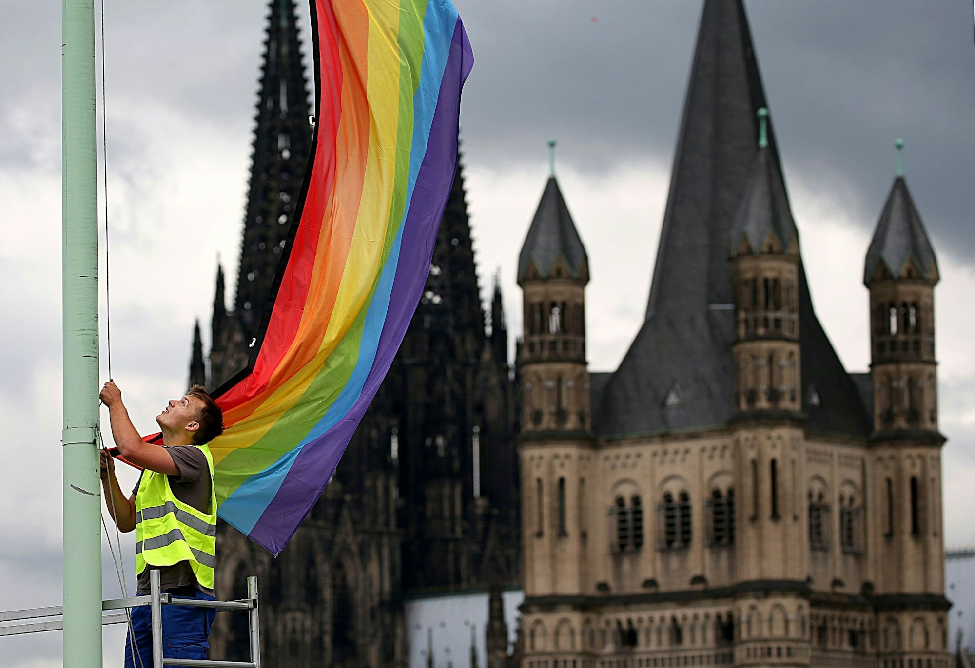 Ein Mann hisst eine Regenbogenfahne vor der Kulisse des Kölner Doms und der romanischen Kirche Groß St. Martin - eben das soll bei der Eröffnung eines erzbischöflichen Bildungszentrums nicht erwünscht gewesen sein (Archivbild).