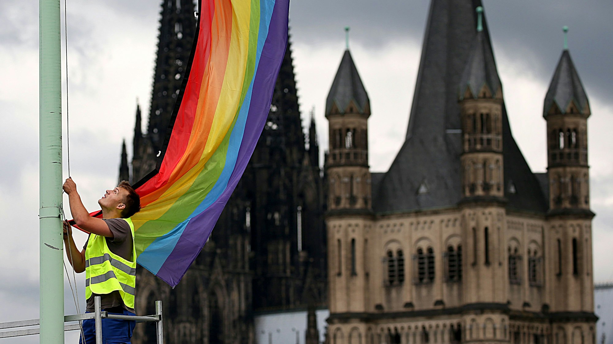 ARCHIV - 30.06.2017, Nordrhein-Westfalen, Köln: Ein Arbeiter hängt nach der Entscheidung im Bundestag über die "Ehe für alle" vor der Kulisse des Doms und der Kirche Groß St. Martin (r) eine Regenbogenfahne auf. (zu dpa: «Konflikt um Regenbogen-Symbol bei kirchlicher Schuleröffnung») Foto: Oliver Berg/dpa +++ dpa-Bildfunk +++