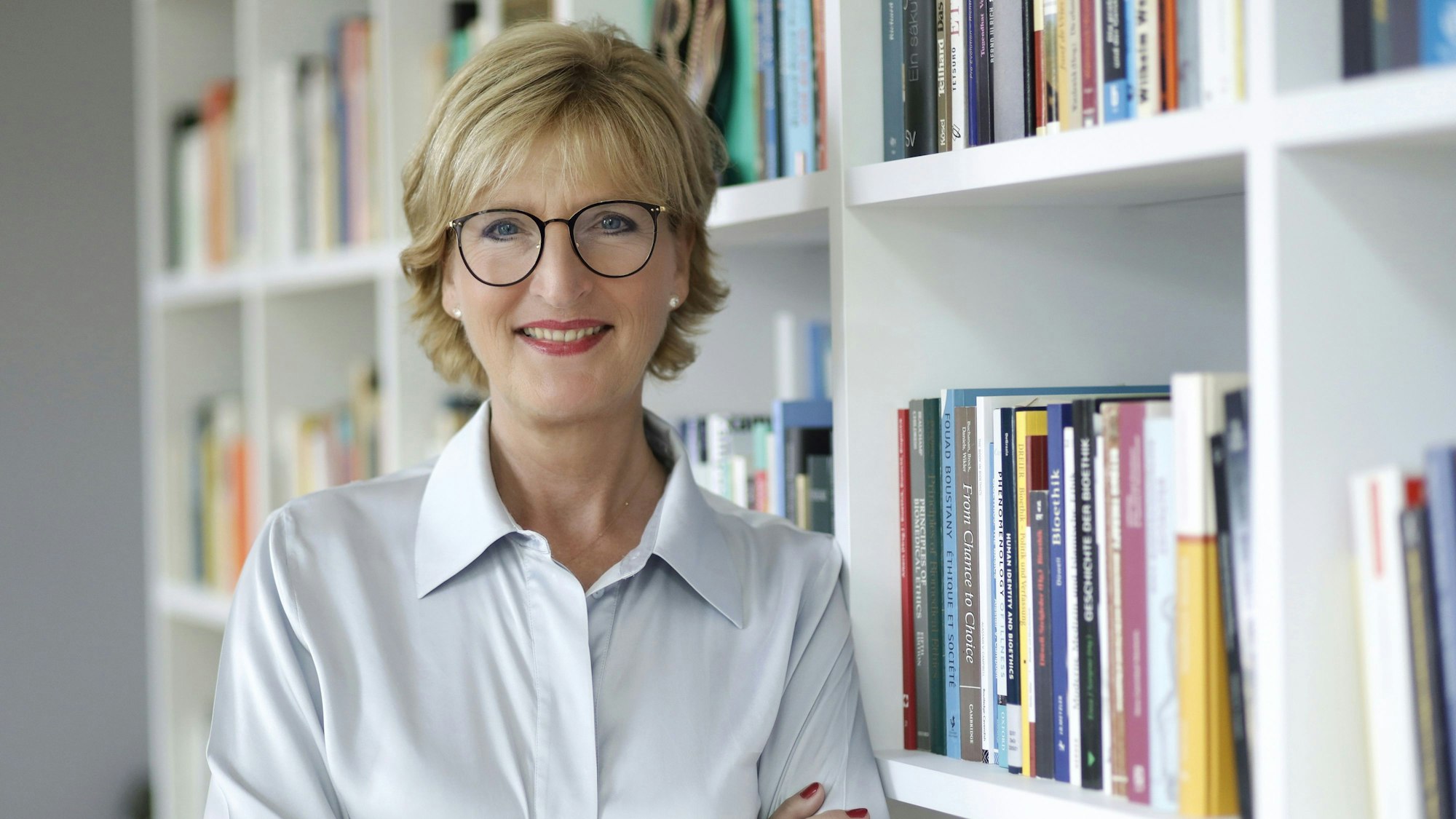 Frau Woopen steht vor einem Bücherregal und lächelt in die Kamera.