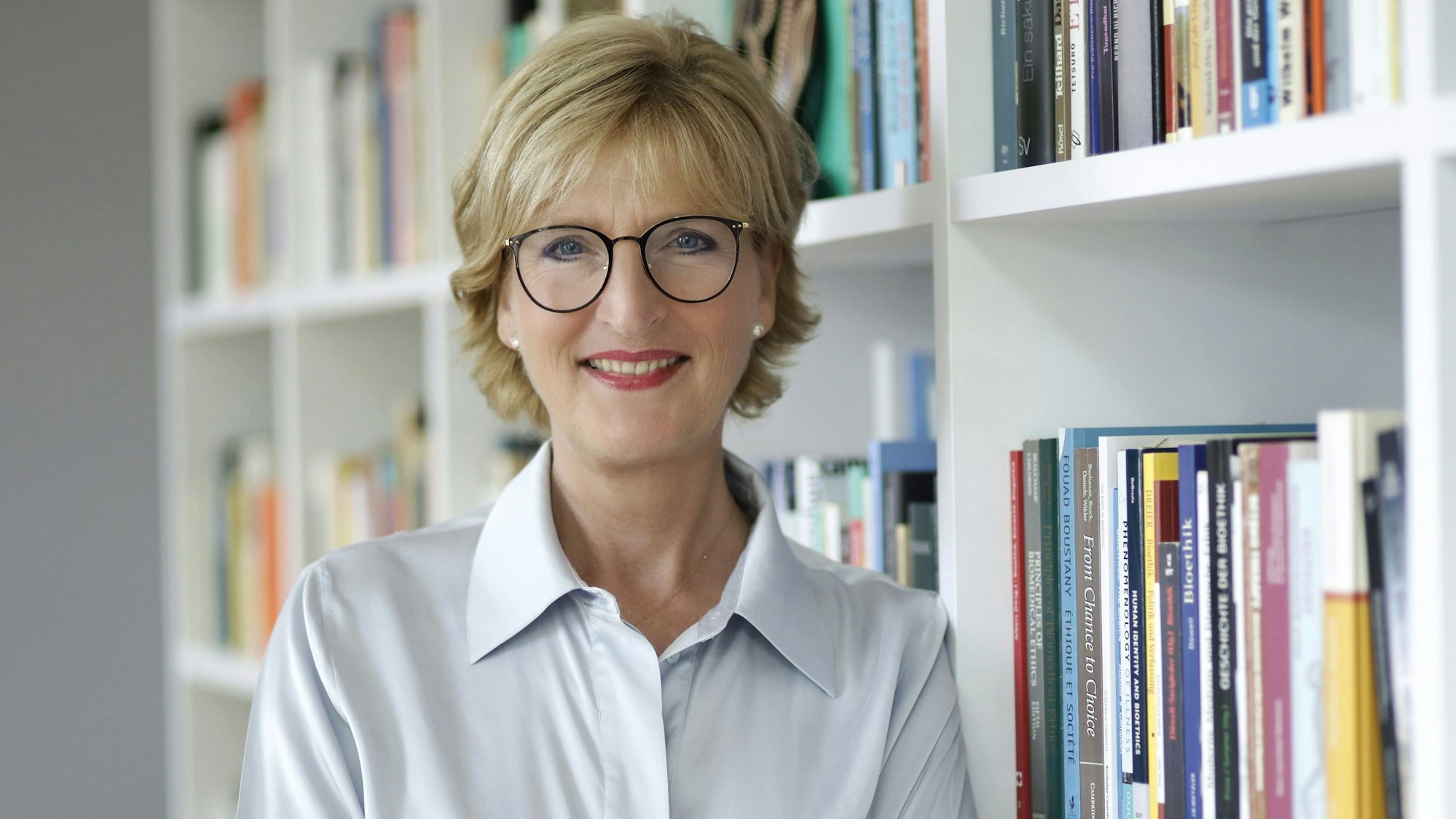 Christiane Woopen steht vor einem Bücherregal und lächelt in die Kamera.