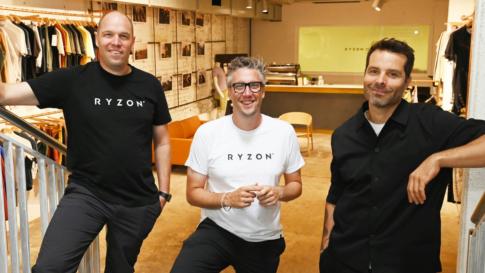 Markus Konrad, Marius Konrad und Fabian Jung stehen im Ryzon-Store im Belgischen Viertel. Im Hintergrund ist der Laden mit ausgestellten Kleidungsstücken und einer Espressobar zu sehen.