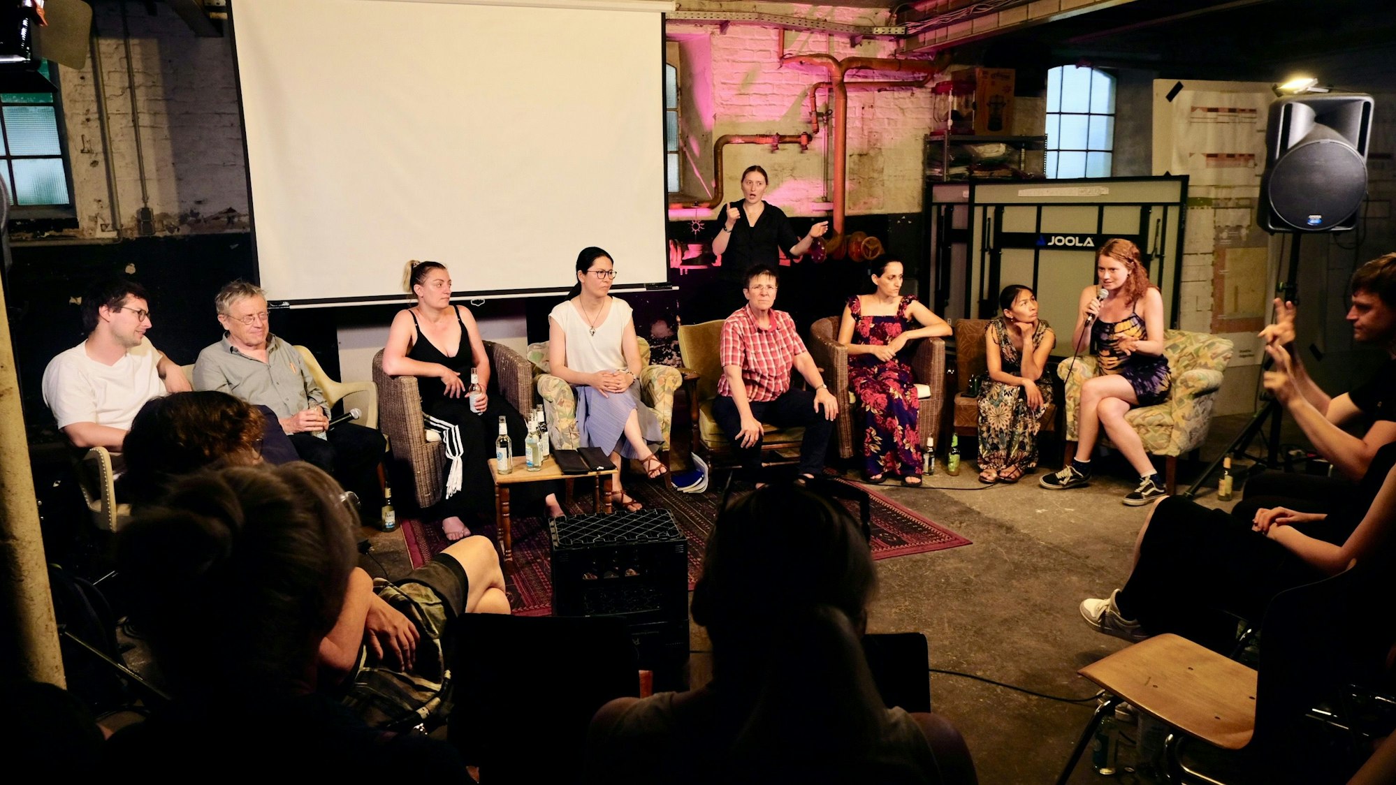 Auf Sesseln in einem großen Raum sitzen Menschen, eine Frau spricht in ein Mikrofon.