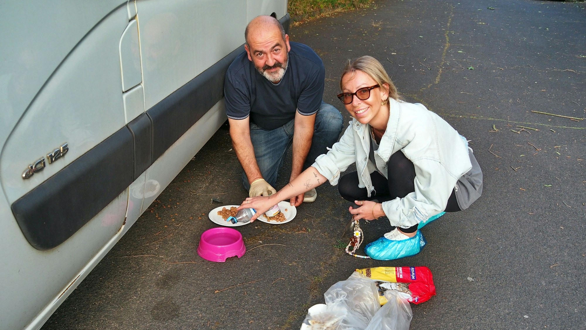 Ein Mann und eine Frau füllen Katzenfutter auf Tellerchen, die Frau füllt Wasser in einen Napf.