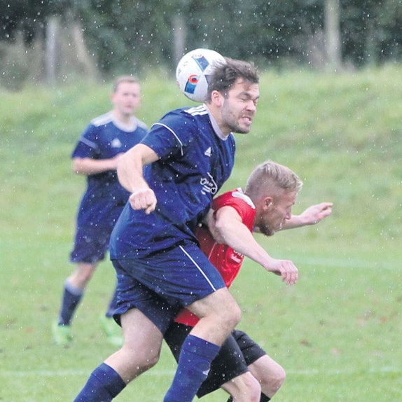 Es regnet auf einem Fußballplatz. Zwei Spieler sind im Zweikampf. Einer der beiden bekommt den Ball gegen den Hinterkopf und hat die Augen geschlossen.