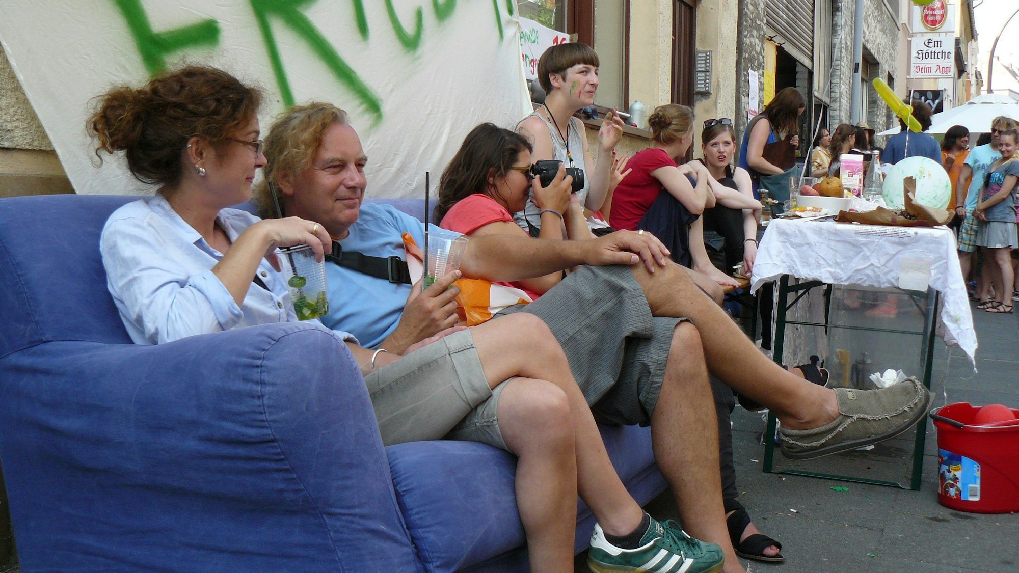Zwei Leute sitzen mit Getränken auf einer Couch am Straßenrand, dahinter ist ein Stand mit Essen und Getränken.