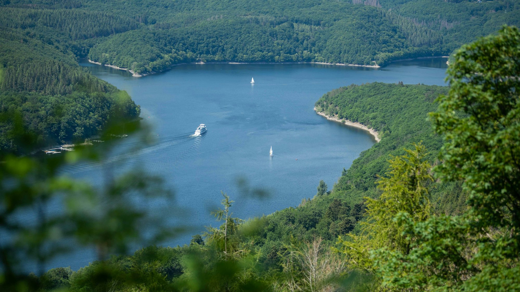 Zwei Segelboote und ein Schiff fahren auf dem Rursee in der Eifel auf dem Wasser.