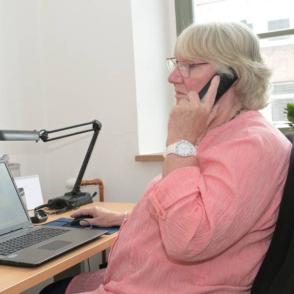 Eine Frau sitzt vor einem Laptop und hält einen Telefonhörer ans Ohr.