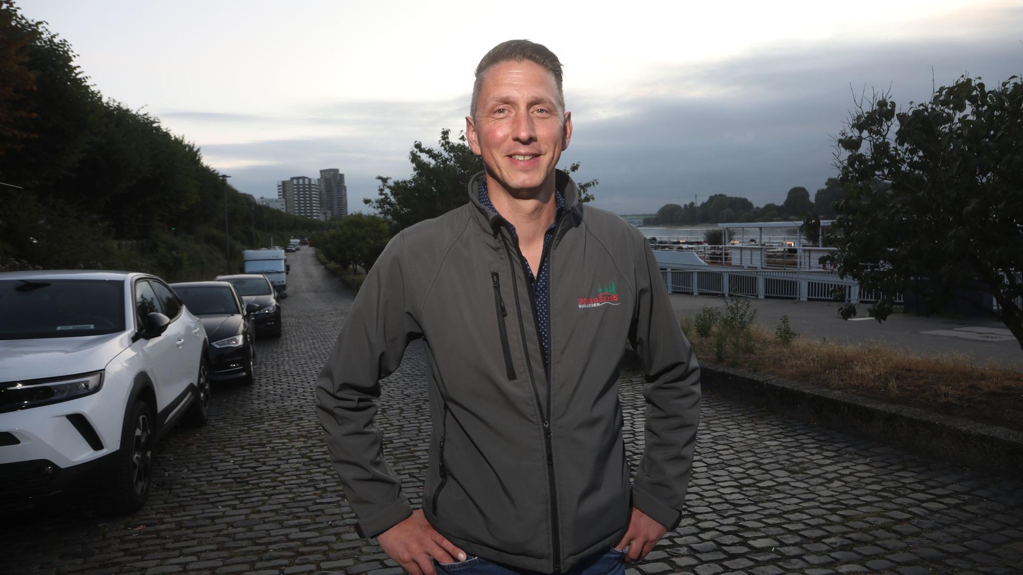 09.07.2025, Köln: Markus Klein hat die Hände an seine Hüfte gestemmt. Er steht auf einer gepflasterten Straße, an der Autos parken.