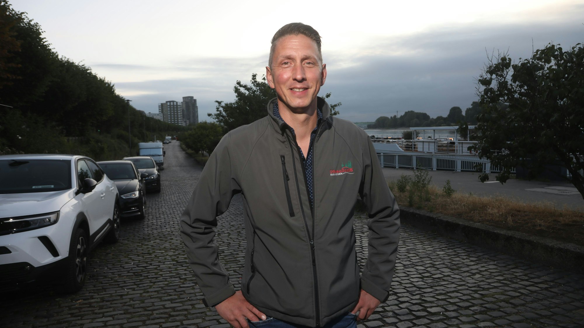 09.07.2025, Köln: Markus Klein hat die Hände an seine Hüfte gestemmt. Er steht auf einer gepflasterten Straße, an der Autos parken.