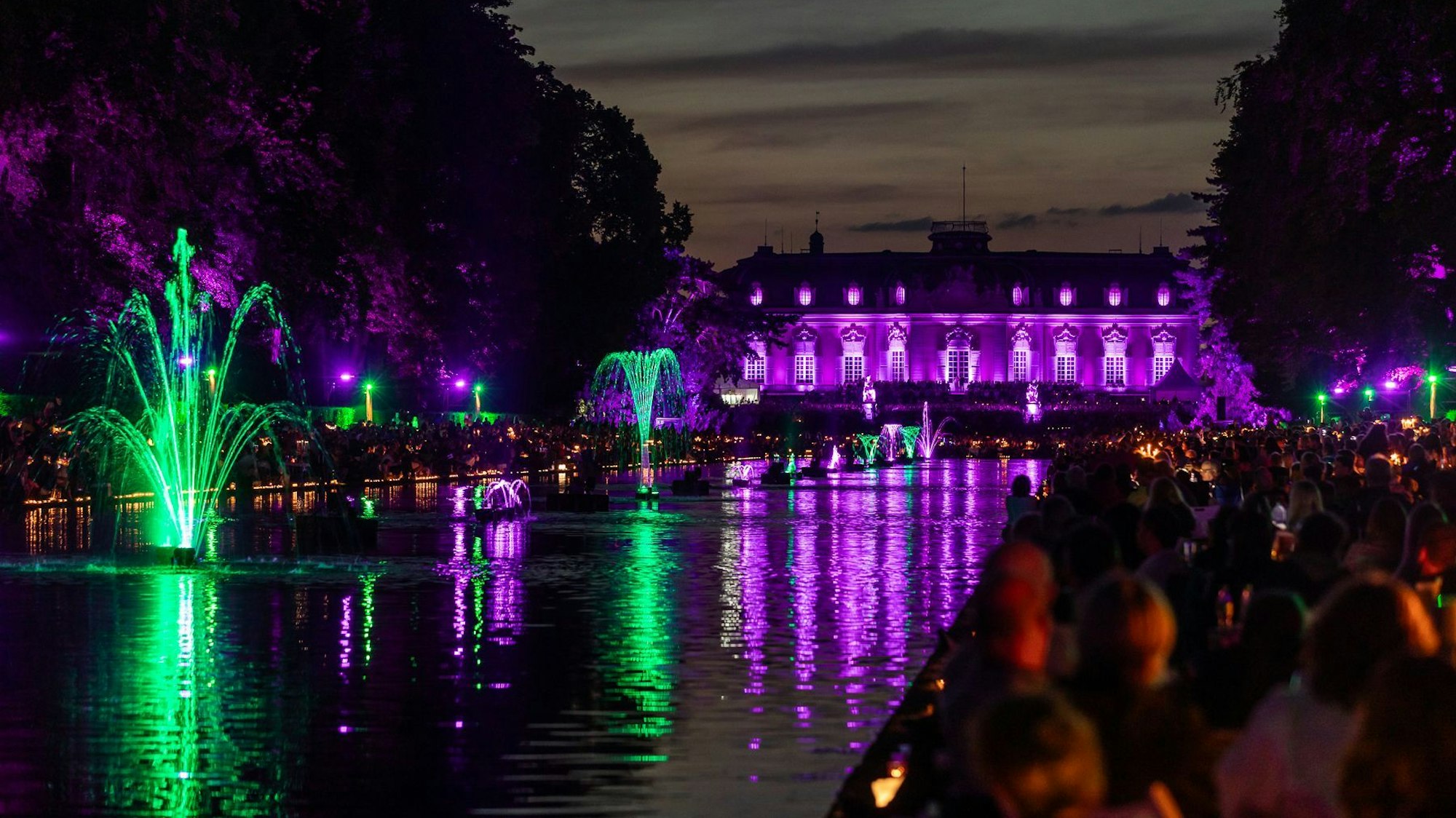 Ein bunt beleuchtetes Schloss am Wasser, die Fontänen sind ebenfalls beleuchtet.