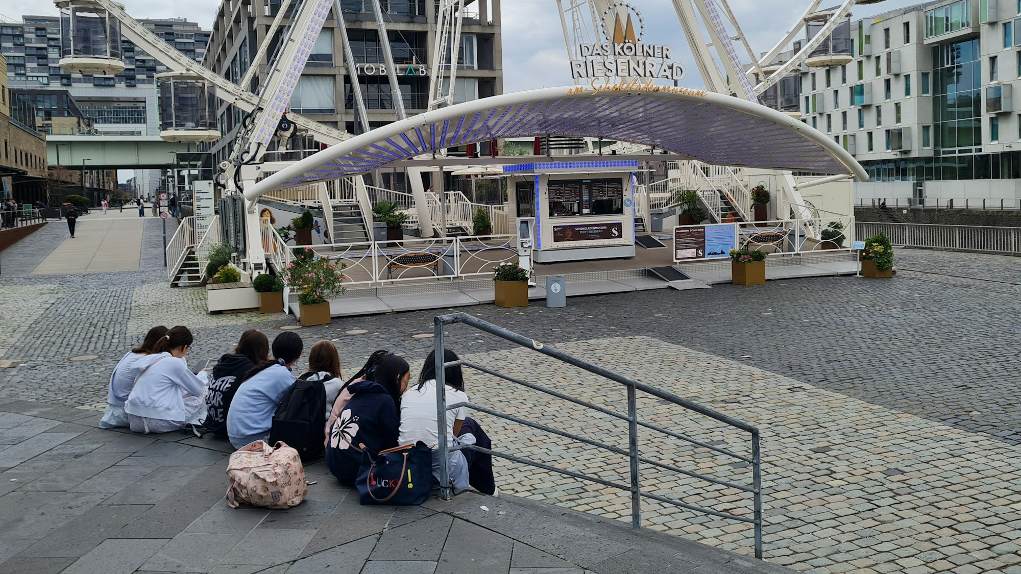 Eine Gruppe Kinder sitzt auf den Treppenstufen zwischen Schokoladenmuseum und Riesenrad.