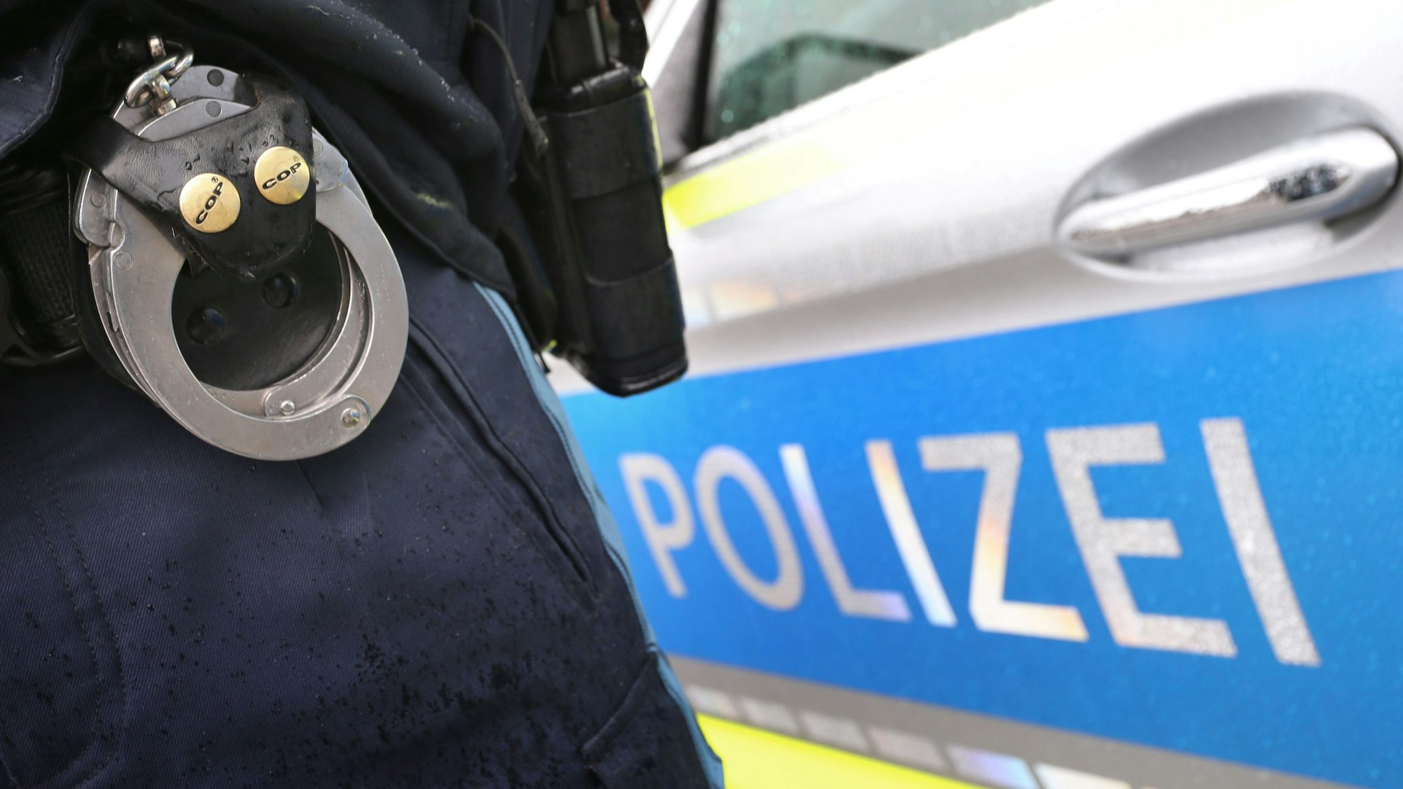 Ein Polizist mit Handschellen und einer Pistole am Gürtel steht vor einem Streifenwagen.