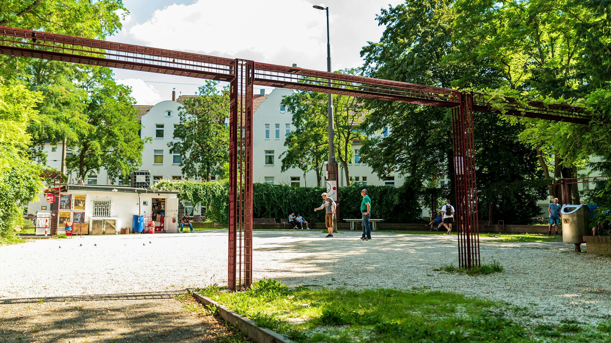 Ein kleiner Platz mit Kiosk und Tischtennisplatte im Hintergrund, im Vordergrund spielen zwei Menschen Boule.