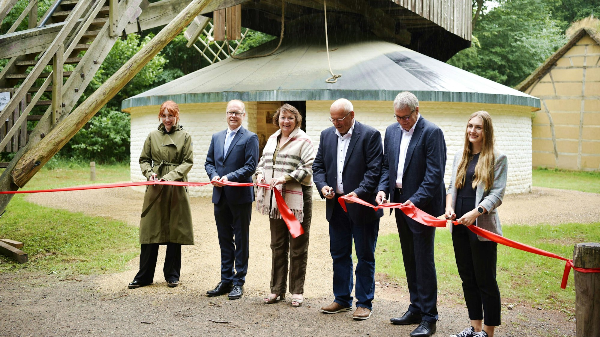 Drei Frauen und drei Männer stehen vor einem weißen, gemauerten Gebäude und schneiden ein rotes Band durch.