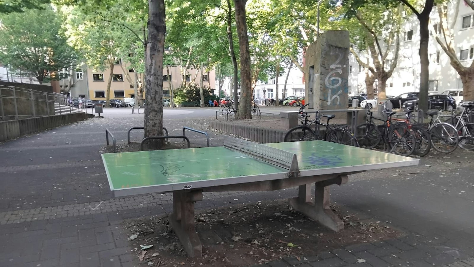 Eine Tischtennisplatte auf einem Platz, Bäume und Fahrräder im Hintergrund.