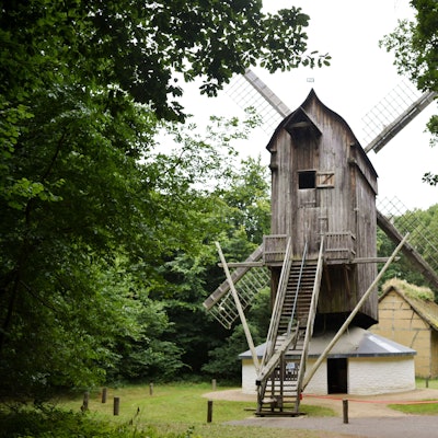 Eine Bockwindmühle aus Holz steht zwischen grünen Bäumen.