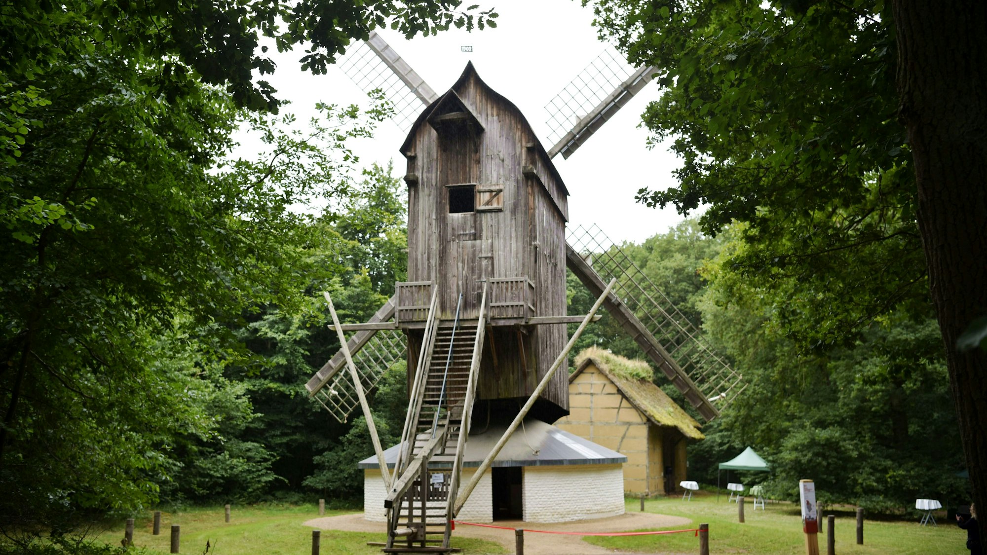 Eine Bockwindmühle aus Holz steht zwischen grünen Bäumen.