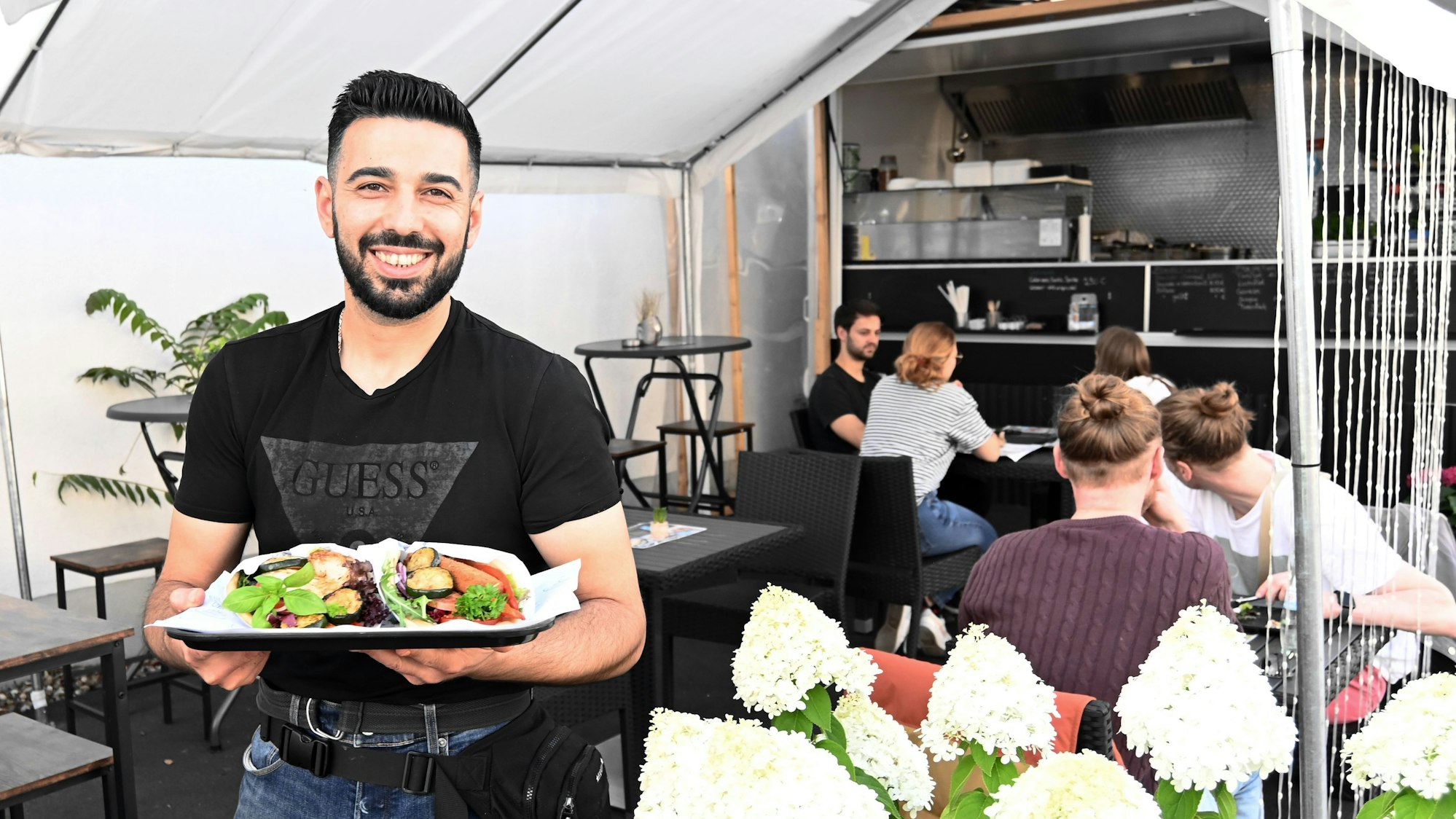Der Koch zeigt im Freisitz einen Pita-Teller und lacht in die Kamera.