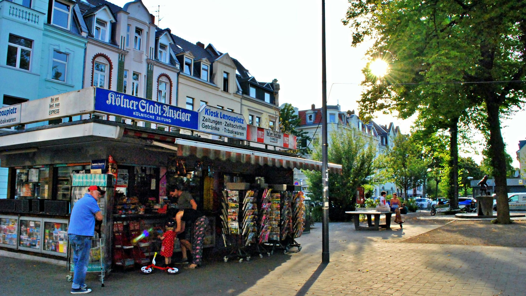 Ein Platz in der Abendsonne, im Vordergrund ein Kiosk, im Hintergrund eine Tischtennisplatte, auf der zwei Leute sitzen.