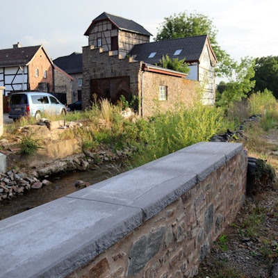 Die Erftmauern in Arloff sind errichtet, in der Mitte fließt der Bach, im Hintergrund stehen Häuser.