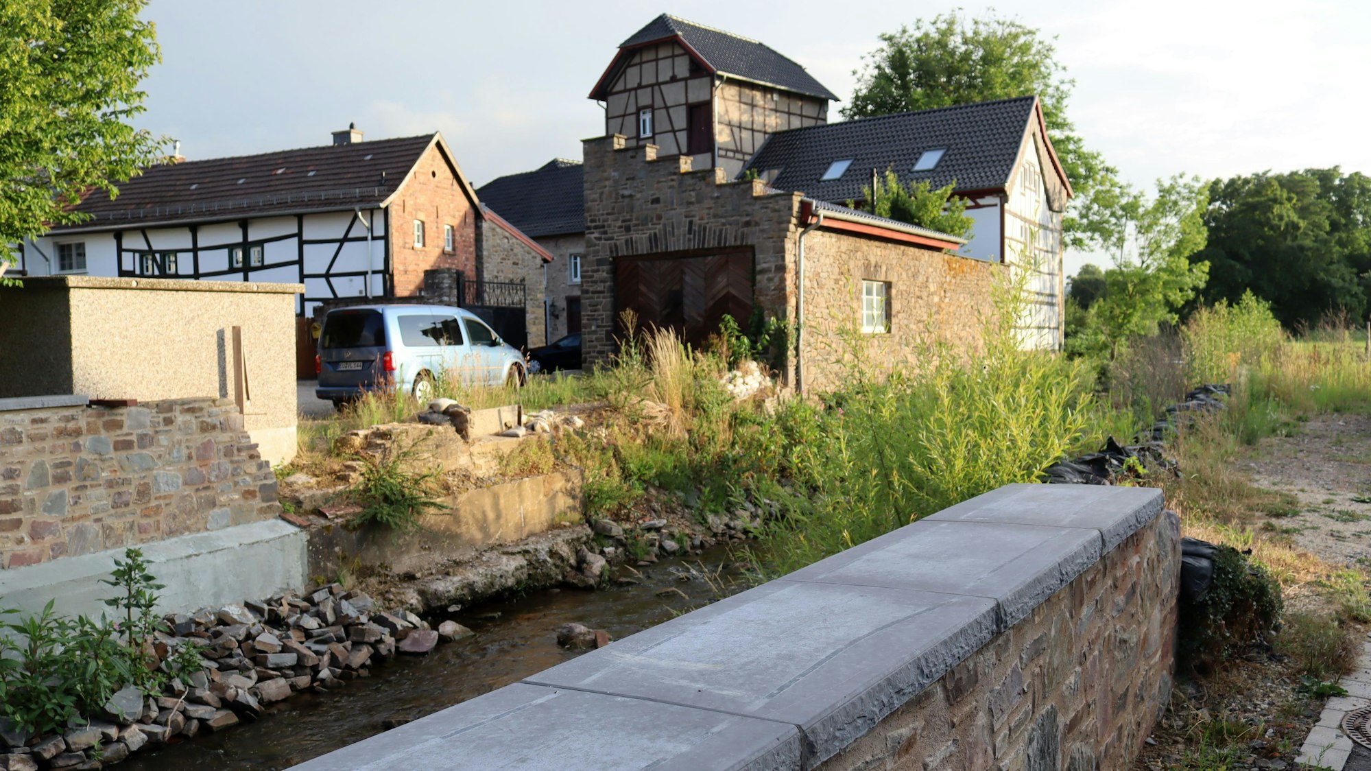 Die Erftmauern in Arloff sind errichtet, in der Mitte fließt der Bach, im Hintergrund stehen Häuser.