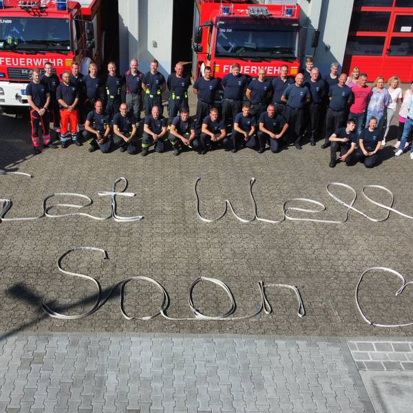 Feuerwehrleute vor einer Fahrzeughalle; auf dem Boden ist der Satz „Get well soon“ zu lesen.
