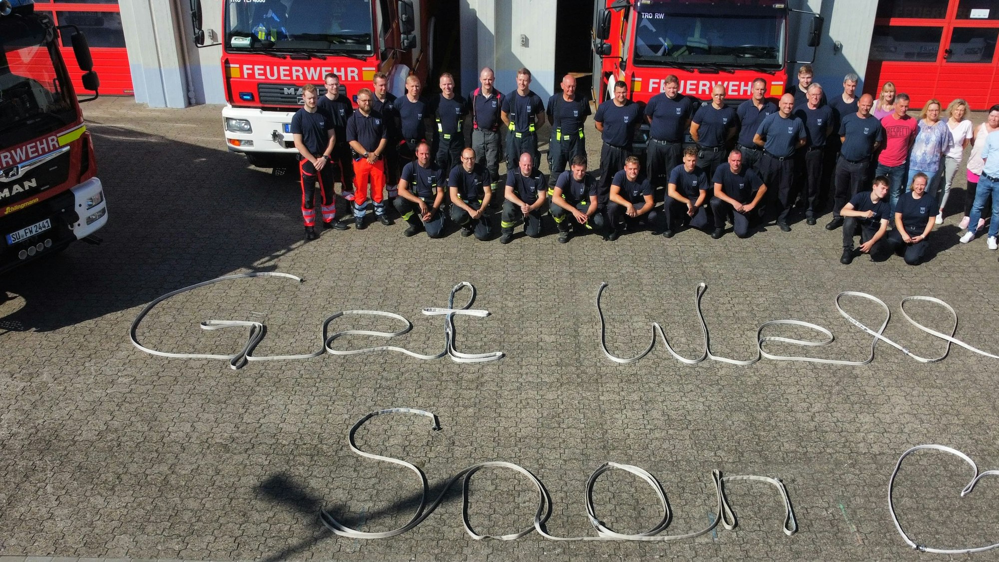 Feuerwehrleute vor einer Fahrzeughalle; auf dem Boden ist der Satz „Get well soon“ zu lesen.