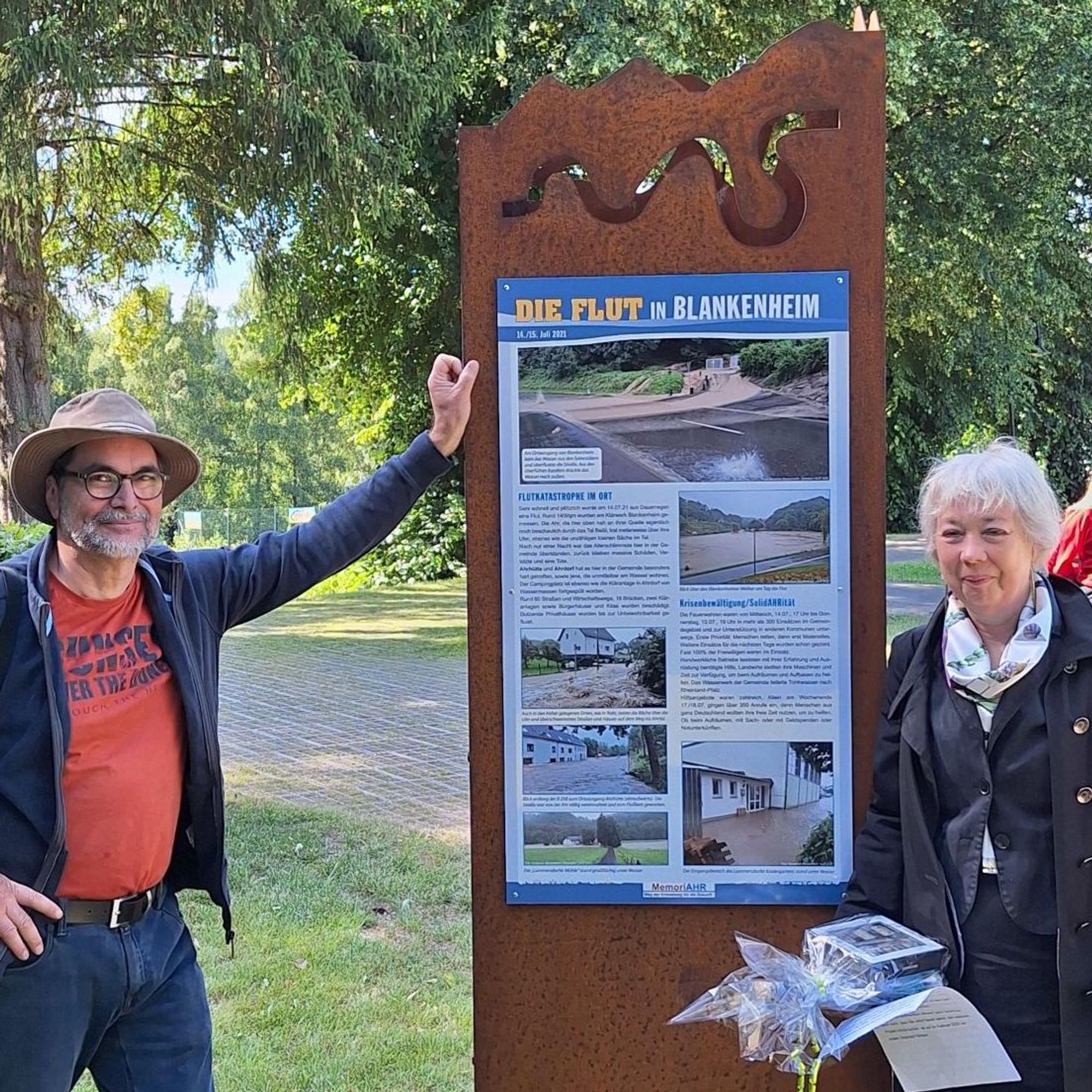 Annette Holzapfel und Andreas Schmickler stehen an einer Stele, auf der „Die Flut in Blankenheim“ steht. Es sind mehrere Bilder zur Flutkatastrophe mit Textelementen versehen.