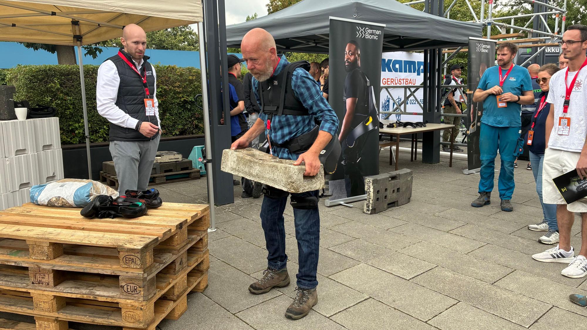 Der Tischler Manfred Kreinjobst kann seinen Beruf nicht mehr ausführen, weil er von Jahrzehnten der Arbeit Hüftprobleme hat. Bei der Handwerkskammer Köln hebt er einen 35 Kilogramm schweren Betonfuß mithilfe eines Exoskeletts mühelos hoch.