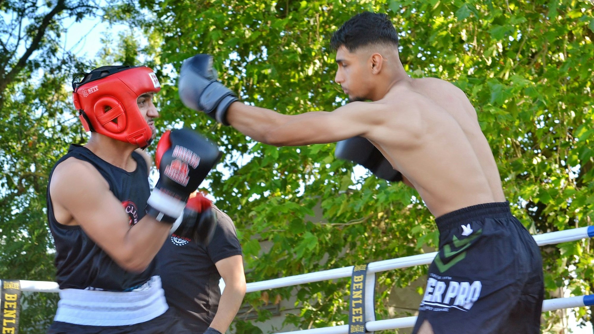 Zwei Jugendliche kämpfen in einem Boxring, der draußen liegt. Einer der beiden schlägt in Richtung des Kopfes des anderen.