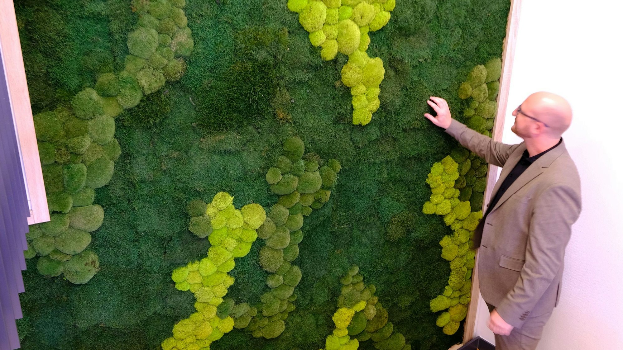 Ein Mann im Anzug berührt eine mit Moos gestaltete Wand in einem Treppenhaus.