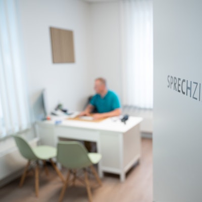 Ein Hausarzt sitzt in einem Sprechzimmer seiner Hausarztpraxis an einem Schreibtisch.