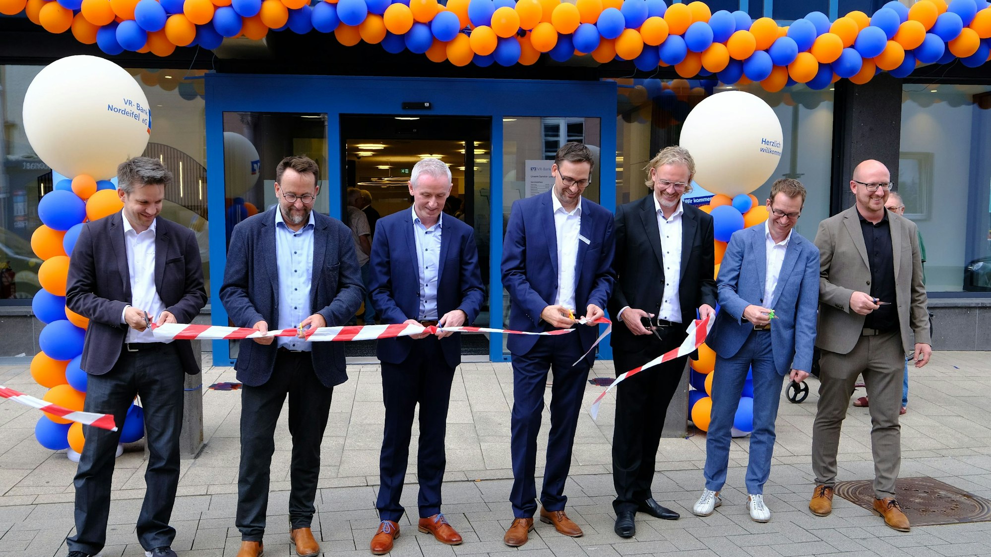 Wiedereröffnung der VR-Bank in Schleiden: Sieben Männer in Anzügen zerschneiden symbolisch ein rot-weißes Flatterband.