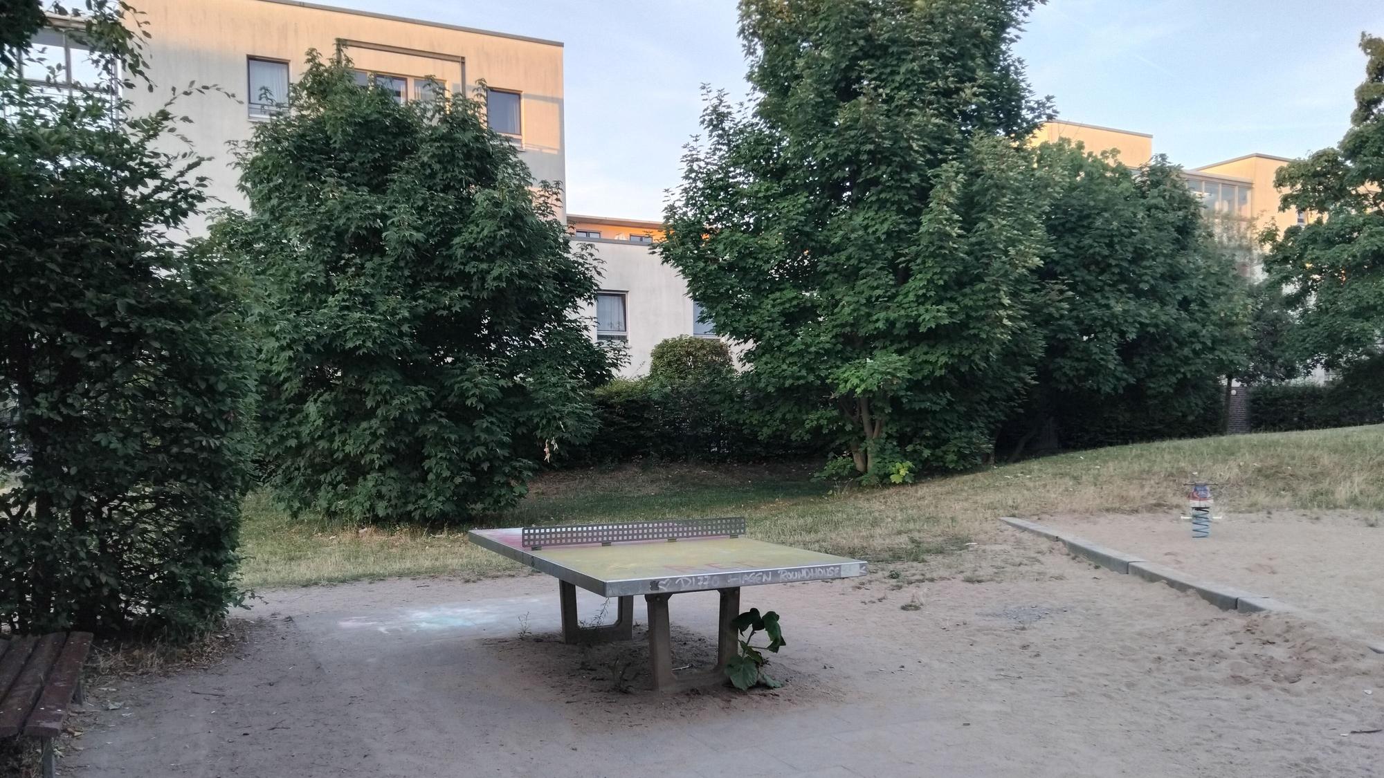 Tischtennisplatte neben einem Spielplatz, im Hintergrund Bäume und Wohngebäude.